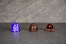 Three chocolate candies are displayed on a gray surface: a wrapped purple sugar-free candy on the left, a solid brown chocolate in the middle, and a half-cut chocolate revealing a red filling on the right.